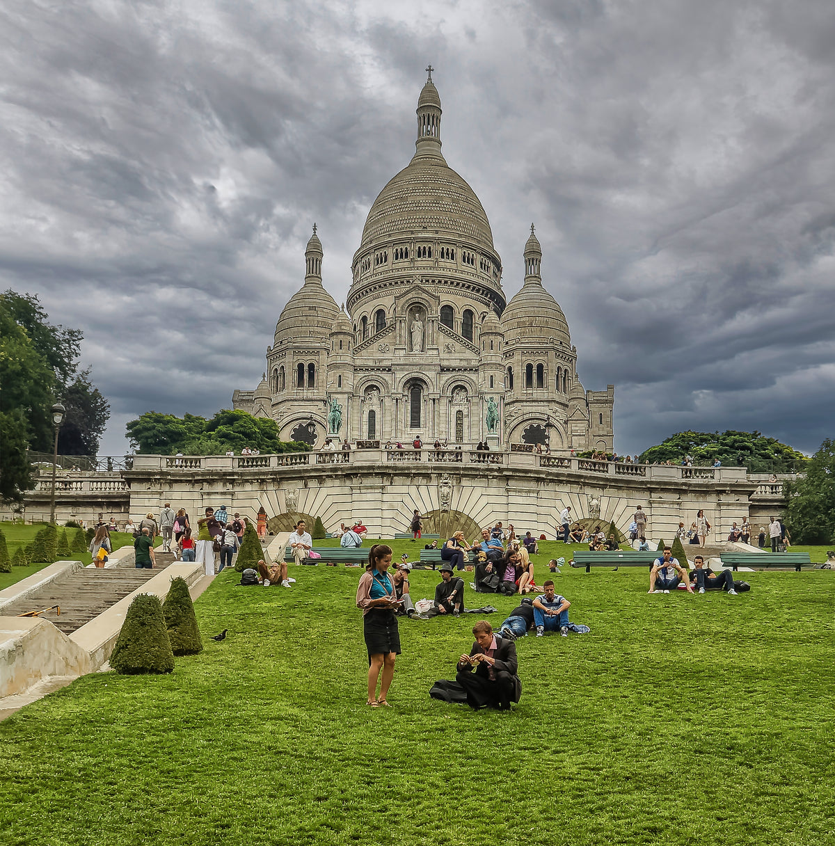 La basílica del Sacré-Cœur en blanco sobre un cielo plomizo, con gente en el césped. Fotografía fine art, edición limitada de 5 ejemplares.