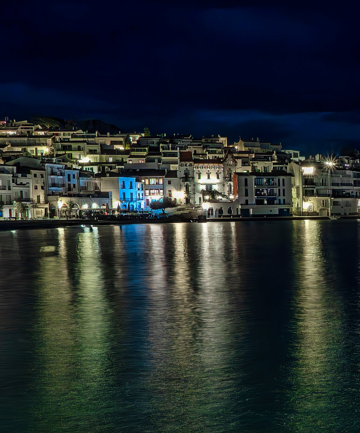 White houses of Cadaqués at night with green and golden reflections on the water. Fine art photograph, limited edition of 5 copies.