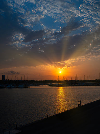Sol bajo con rayos entre las nubes sobre el puerto de Cambrils y una figura en el dique. Fotografía fine art, edición limitada de 5 ejemplares.